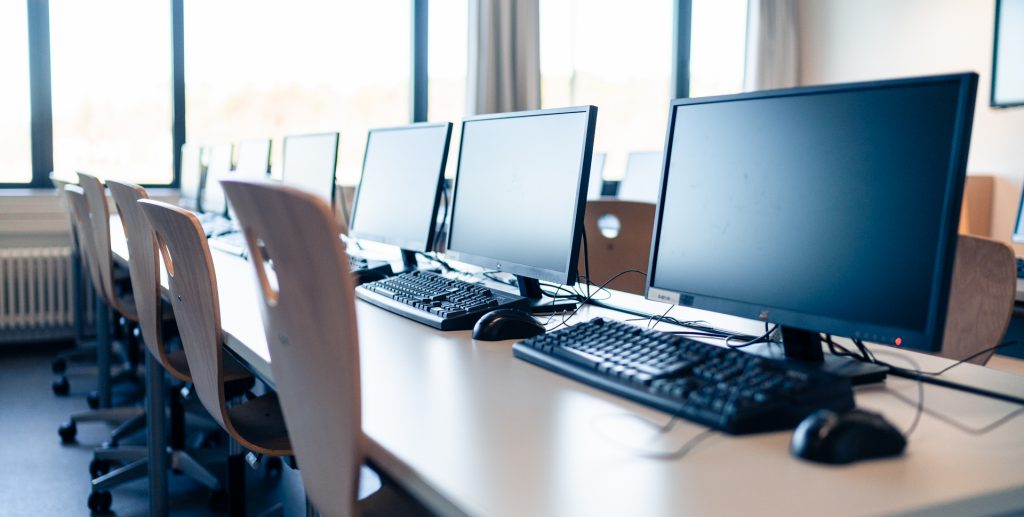 Reihe von Computerarbeitsplätzen mit Monitoren und Tastaturen in einem Klassenraum.