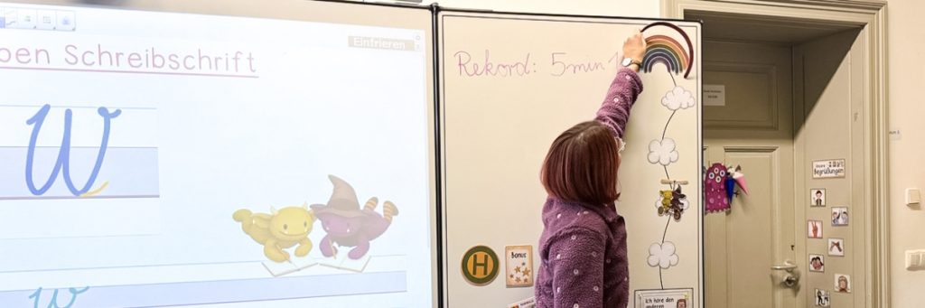 Eine Lehrerin fügt im Klassenzimmer einen Farbbogen eines Regenbogens an ein Tafelbild mit Wolken und einem Tandemrad hinzu, das den Lernfortschritt ihrer Klasse symboliseren soll.