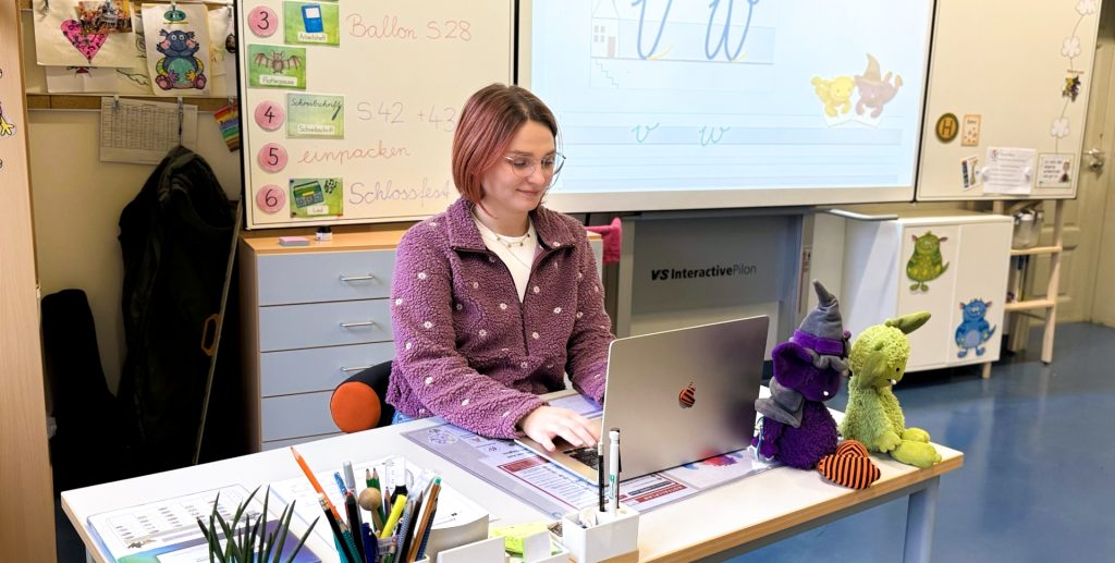 Grundschullehrerin Lena Zscheuschler sitzt in einem Klassenzimmer vor einem Whiteboard am Schreibtisch. Auf dem Whiteboard sind die Schreibschrift-Buchstaben V und W zu sehen und die beiden Kuschelmonster, die auch als Figuren vor ihrem Laptop auf dem Schreibtisch sitzen.