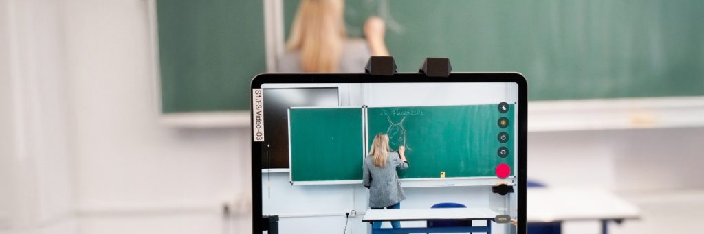 Eine Frau in Jeans und grau-weiß kariertem Jackett mit rotblonden langen Haaren ist von hinten zwei Mal vor einer grünen Schultafel zu sehen. Einmal verschwommen im Hintergrund und auf dem Monitor mit der Kamera, mit der sie gefilmt wird. Sie hat eine Zelle mit Kreide an die Tafel gemalt und mit der Überschrift "Die Pflanzenzelle" versehen.