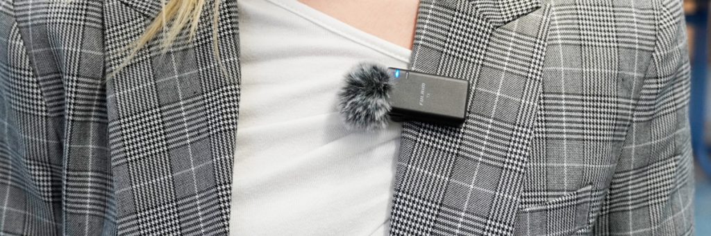 Es ist der Brustbereich einer Frau in einem grau-weiß karierten Jacket und einem hellen T-Shirt zu sehen. Am Revers der Frau klemmt ein kleines Mikro mit Windpuschel.