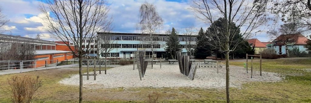 Im Hintergrund ist die türkisblaue Fassade des Schulgebäudes der Bruno-Bürgel-Oberschule vor blauem Himmel zu sehen, im Vordergrund stehen unbelaubte Bäume, und im Mittelbereich ist ein Turn- oder Bolzplatz mit Holzgeräten zu erkennen.
