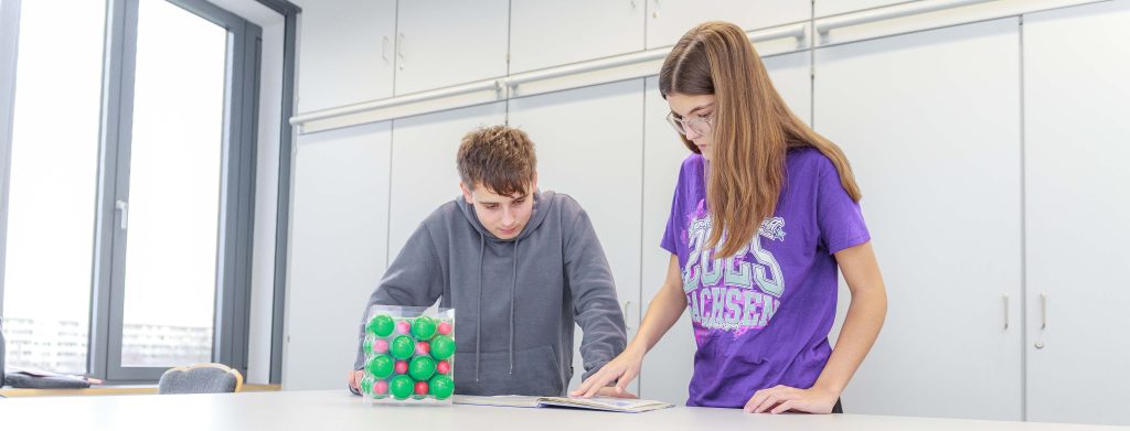 Zwei Schüler, ein Junge und ein Mädchen, stehen an einem Tisch und betrachten ein Buch. Auf dem Tisch liegt ein Behälter mit bunten Bällen.