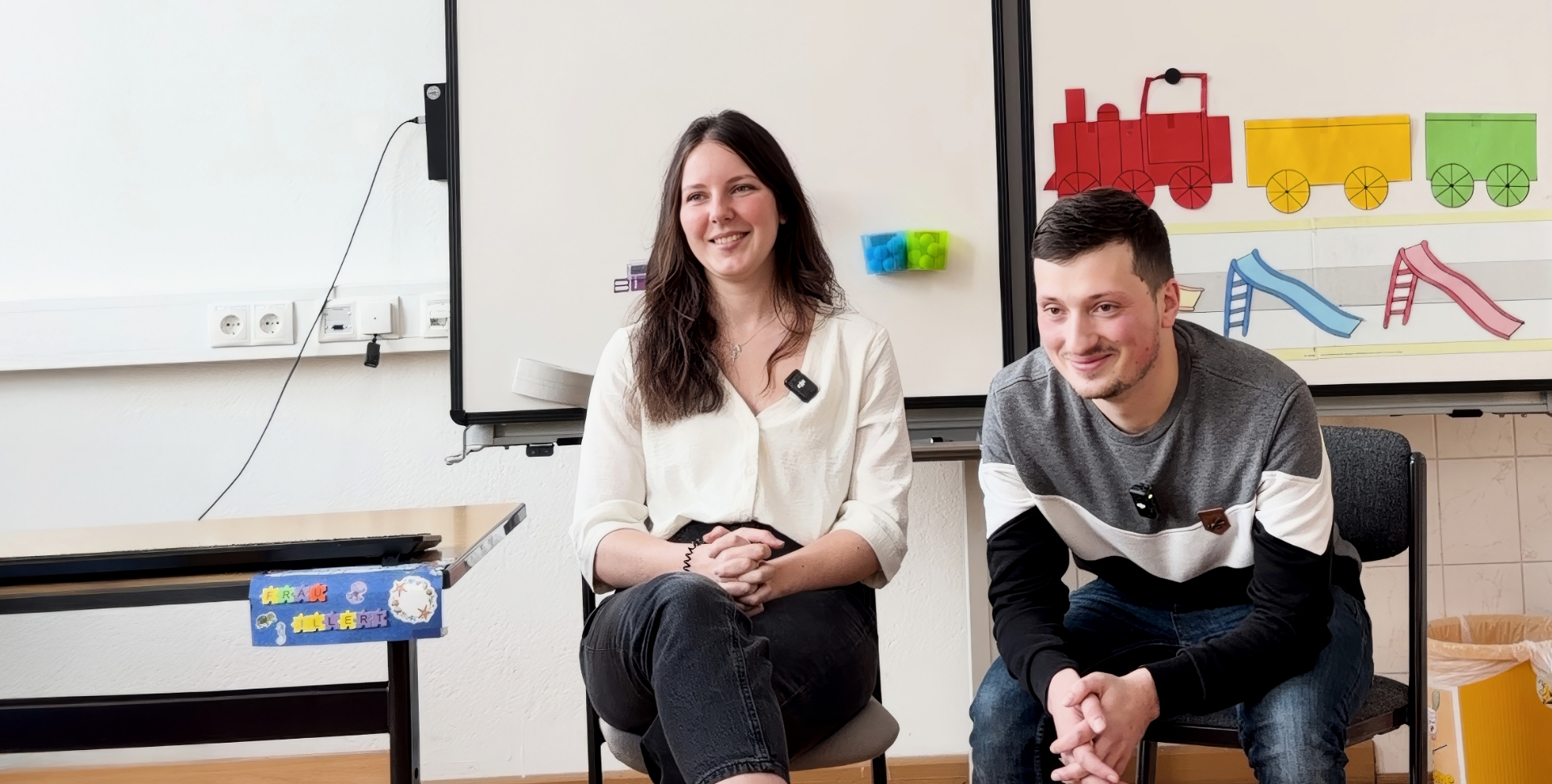 Eine Frau und ein Mann sitzen in einem Klassenzimmer vor einer weißen Tafel, auf der ein rot-gelb-grüner Güterzug aufgeklebt ist und drei verschiedenfarbige Rutschen darunter.