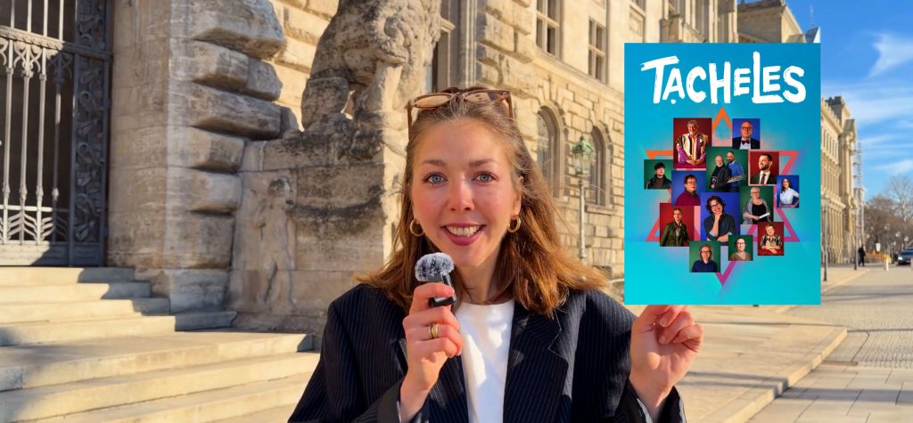Junge Frau hält ein Plakat mit dem Titel 'TACHELES' und verschiedenen Porträts in der Hand, während sie vor einem historischen Gebäude steht.