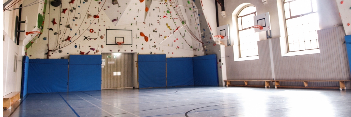 Eine Turnhalle mit Kletterwand. Von der Decke hängen Kletterseile und an der Wand lehnen dicke Sprungmatten.