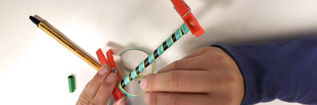 Es sind zwei Kinderhände zu sehen, die eine auf einen Stift gerollte Luftschlange mit einer Botschaft für eine Skytale beschriften. Eine Skytale ist eine seit der antike bekannte mechanische Verschlüsselungstechnik, die auf diesem Bild eine Übungsaufgabe im Informatikunterricht illustriert.