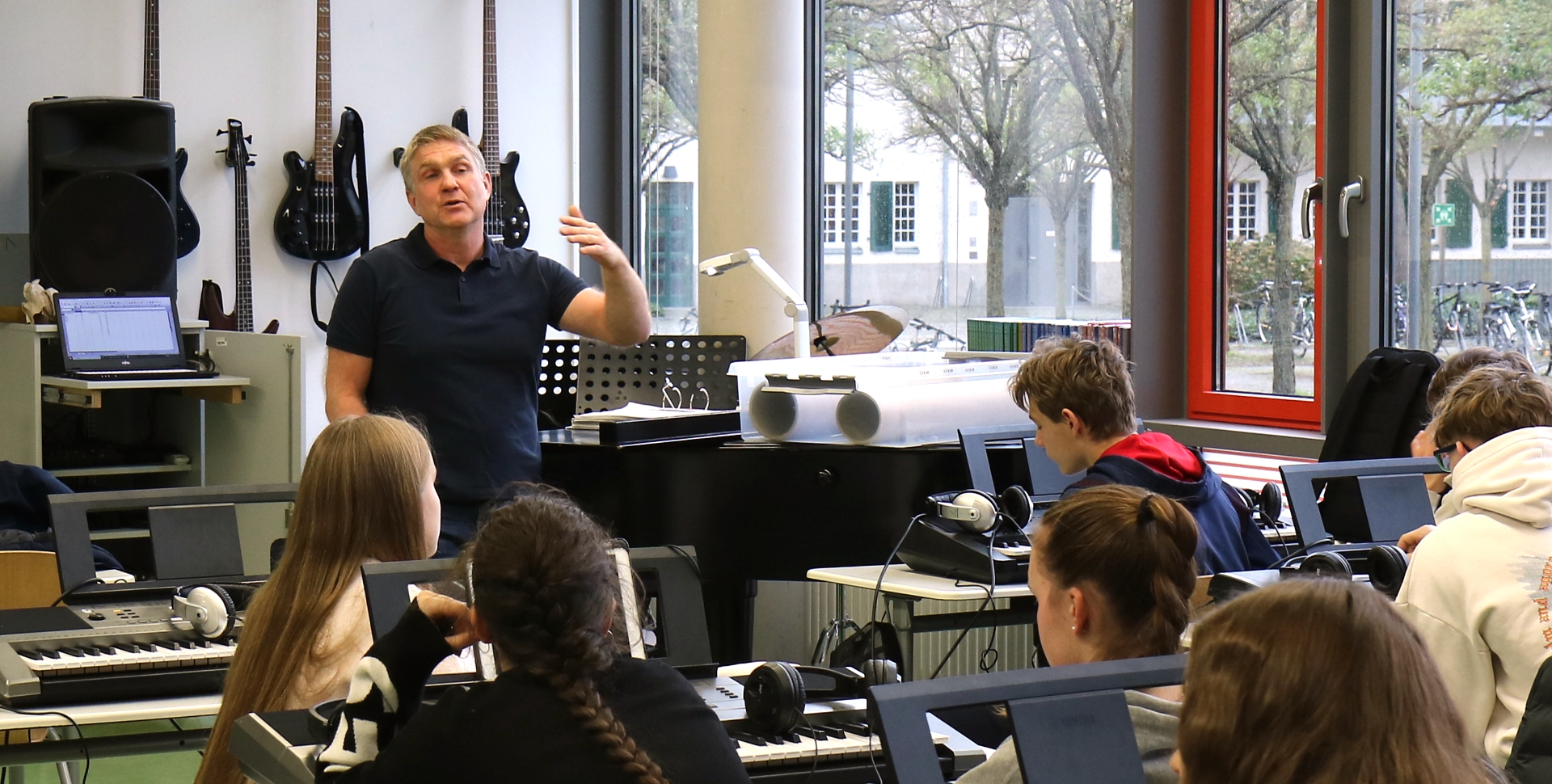 Musiklehrer Maik Wagner steht vor einer Klasse des Sportgymnasiums Dresden und erklärt mit ausholender Armbewegung etwas. Im Hintergrund ist eine Wand mit aufgehängten E-Gitarren zu sehen.