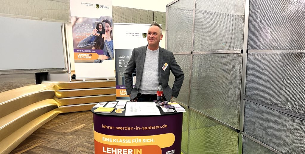 Oliver Damm steht an einem halbrunden Messestand auf einer Bildungsmesse. Der Stand ist mit einem Banner von "Lehrer werden in Sachsen" beklebt und macht dafür Werbung.