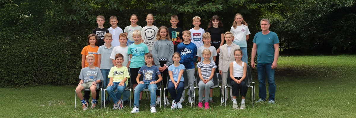 In 3 Reihen aufgestellt und auf Stühlen vorn sitzend sind die Schülerinnen und Schüler der Klasse 5-1 von Klassenlehrer Maik Wagner mit ihm auf dem Klassenfoto vor einer Hecke zu sehen.