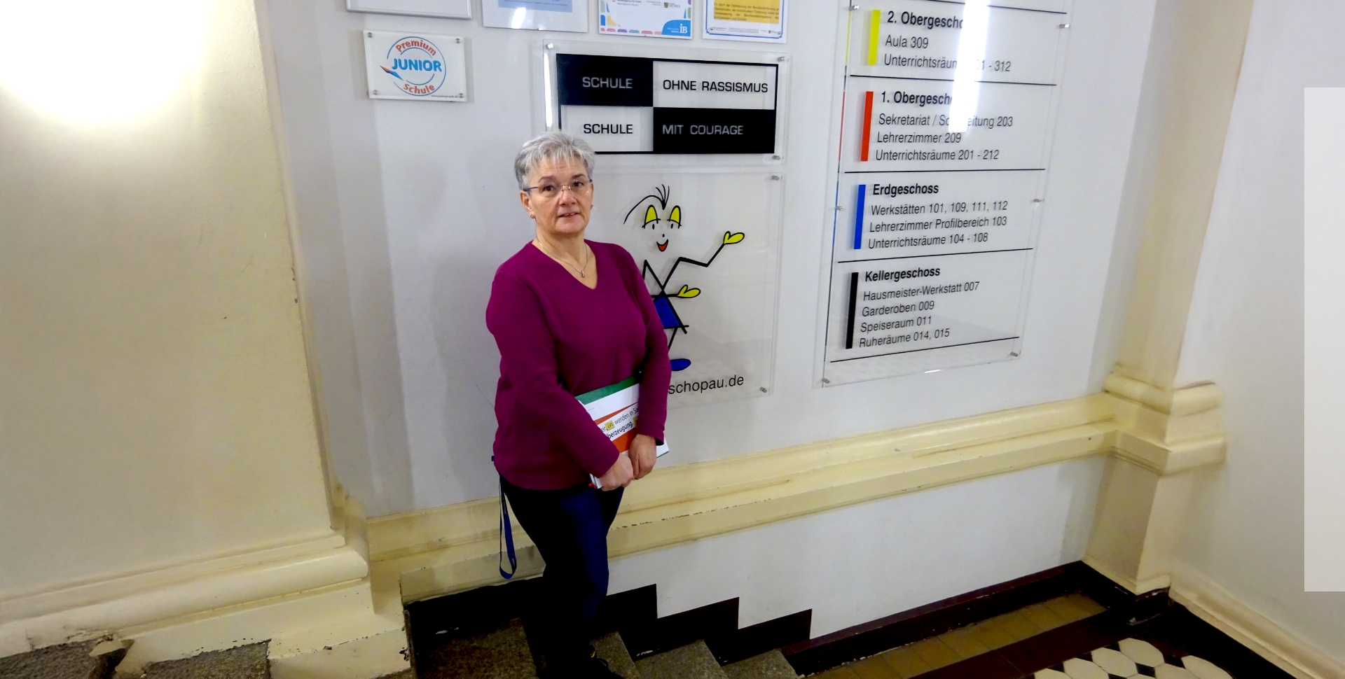 Sybille Höfer, Schulleiterin der Martin-Andersen-Nexö-Oberschule in Zschopau steht im Schulflur vor einer Info-Wand mit Wegweisern auf einer Treppe.