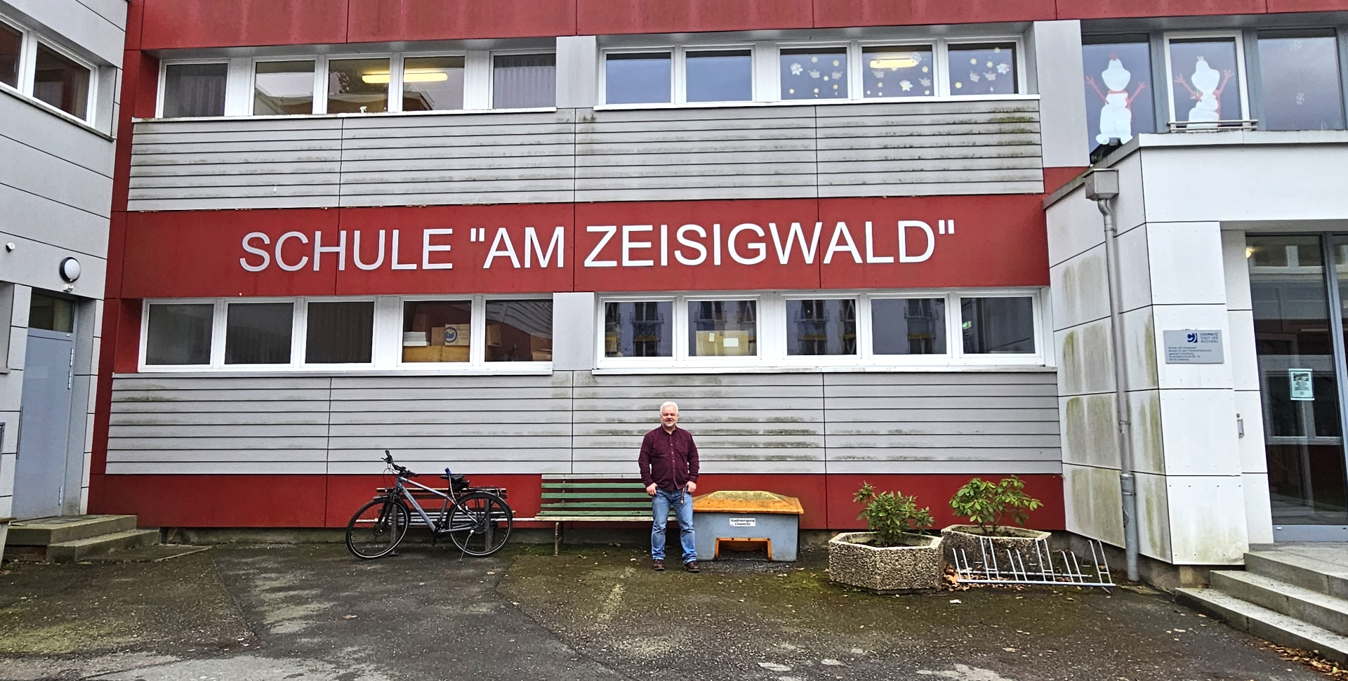 Schulleiter Udo Große steht vor der grau-rot gestreiften Fassade der "Schule am Zeisigwald". Der Schulname ist auf einem roten Farbband an der Fassade angeschrieben.