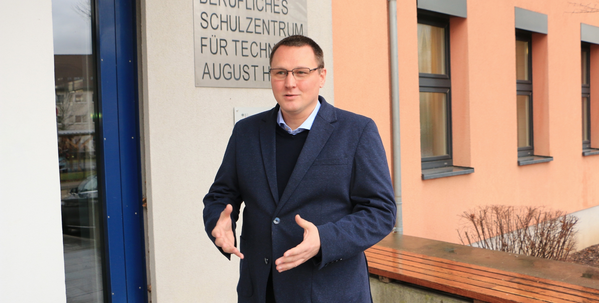 Lutz Langer, Schulleiter des Beruflichen Schulzentrums August Horch in Zwickau, steht vor einem Schild mit dem Namen der Schule und macht eine armöffnende Geste, als ob er im Gespräch etwas erklärt. Im Hintergrund ist eine apricotfarbene Wand mit Fenstern der Schule zu sehen.