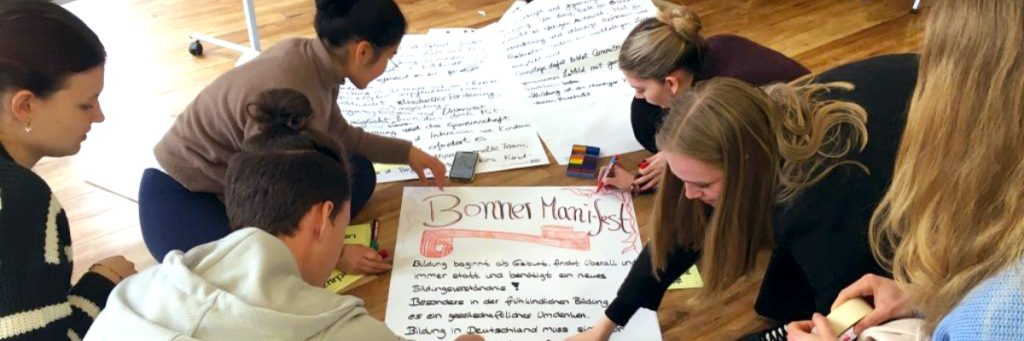 Auf einem Holzboden liegen vor einer Gruppe von sechs jungen Menschen mehrere Flipchart-Bögen. Auf dem vorderen Bogen ist die Überschrift "Bonner Manifest" deutlich erkennbar.