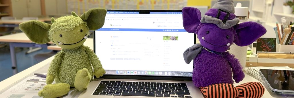 Neben einem aufgeklappten Notebook sitzen rechts ein grünes Kuscheltier-Monster und links ein lilafarbenes.