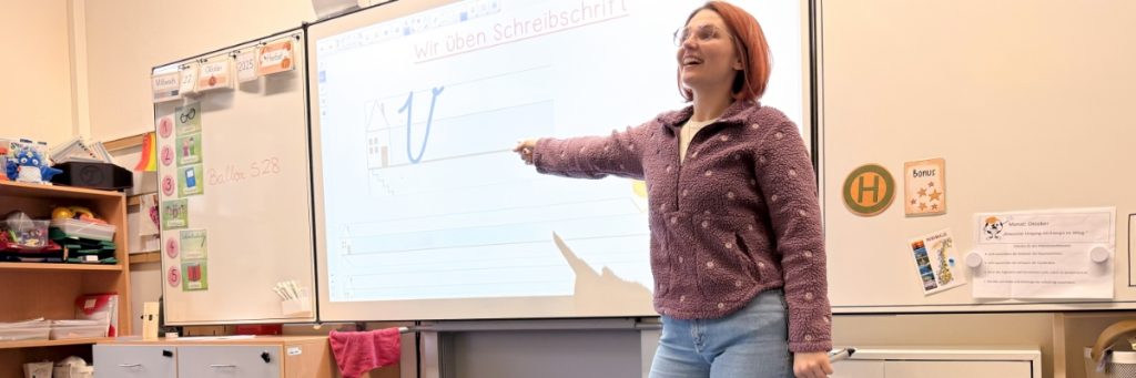 Ein Whiteboard in einem Grundschulklassenzimmer zeigt ein gemaltes Haus und ein ebenso großes V in Schreibschrift. Darüber die Überschrift: Wir üben Schreibschrift. Rechts steht Grundschullehrerin Lena Zscheuschler lächelnd vor dem Board und weist auf die Linien.