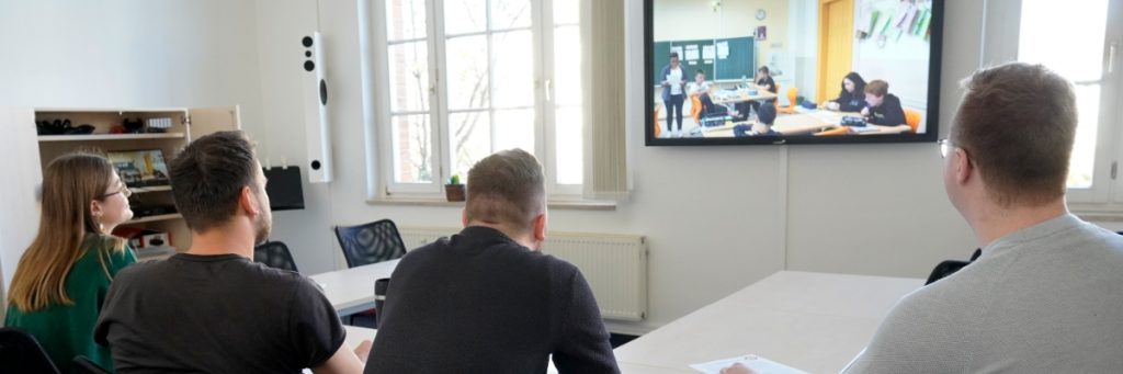 Drei Männer und eine Frau sitzen in einem Raum vor einem Monitor, auf dem eine Schulsituation mit einer Lehrerin und einer Klasse zu sehen ist.