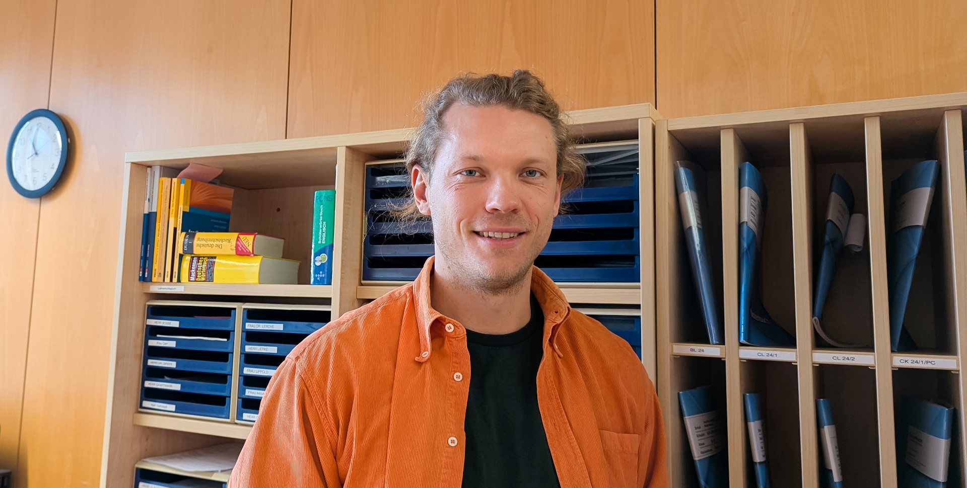 Ein Mann in orangefarbenem Hemd steht in einem Lehrerzimmer vor einer Wand mit Ablagefächern und Notenmappen.