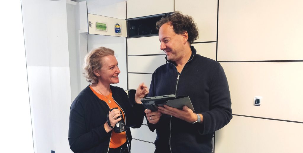 Lehrkräfteausbilderin Anja Urbansky steht links vor einem geöffneten Schließfach und spricht mit ihrem Kollegen Marco Fiedler von der Projektgruppe Videografie, der ein Tablet in seinen Händen hält.