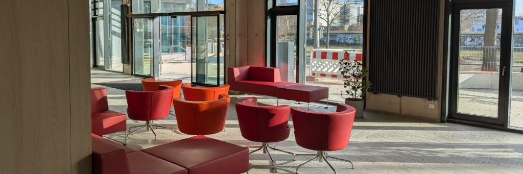 In einer Halle sind mehrere große rote Sessel und ein rotes Sofa vor Glastüren und bodentiefen Fenstern am BSZ Wirtschaft Franz Ludwig Gehe in Dresden aufgestellt. Der Blick fällt auf den noch nicht fertig gestalteten, sonnigen Schulhof mit Baustellenabsperrung und Zäunen im Hintergrund.