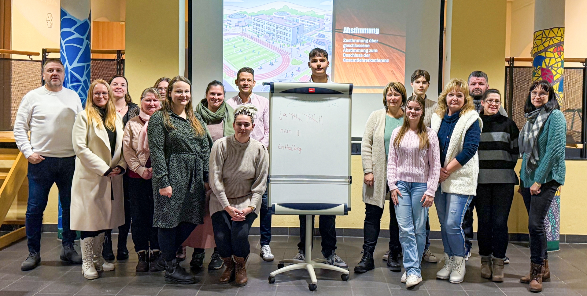 Eine Gruppe von Lehrkräften steht um ein Flipchart, auf dem die Ergebnisse einer Abstimmung zu notiert sind. Im Hintergrund zeigt ein Whiteboard die Zeichnung eines Schulgebäudes sowie den Text "Zustimmung über geschlossene Abstimmung zum Beschluss der Gesamtlehrerkonferenz"
