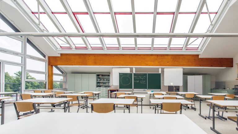 Klassenzimmer mit Tischen, Stühlen und einer Tafel, große Fenster sorgen für viel Tageslicht.