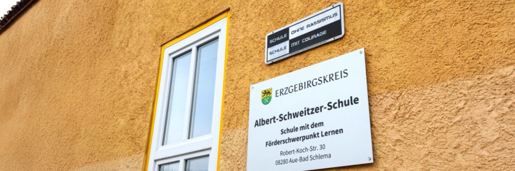 Tafel der Albert-Schweitzer-Schule mit Förderschwerpunkt Lernen an einer gelb verputzten Fassade. Darüber ein weiteres Schild mit der Aufschrift: Schule ohne Rassismus, Schule mit Courage