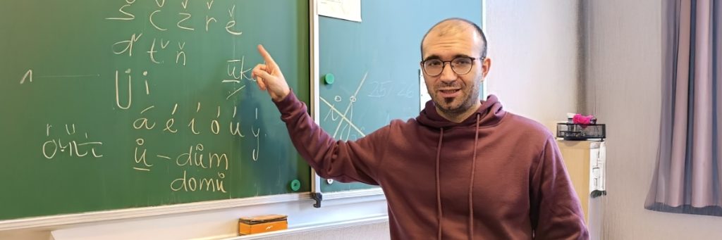 Fremdsprachenassistent Jakub Rudolf steht rechts an einer Tafel und zeigt auf tschechische Buchstaben, die mit gelber Kreide darauf geschrieben stehen.