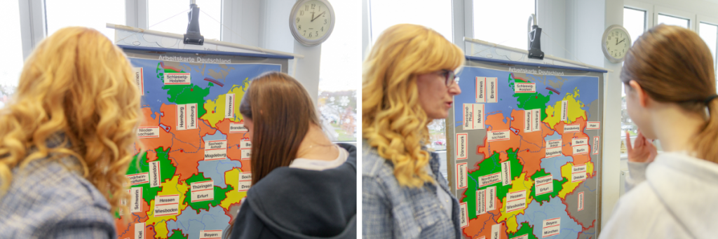 Eine Lehrerin steht links vor einer Deutschlandkarte, auf der die einzelnen Bundesländer mit ihren Hauptstädten auf Stickern beklebt sind. Rechts schaut eine Schülerin ebenfalls auf die Karte.