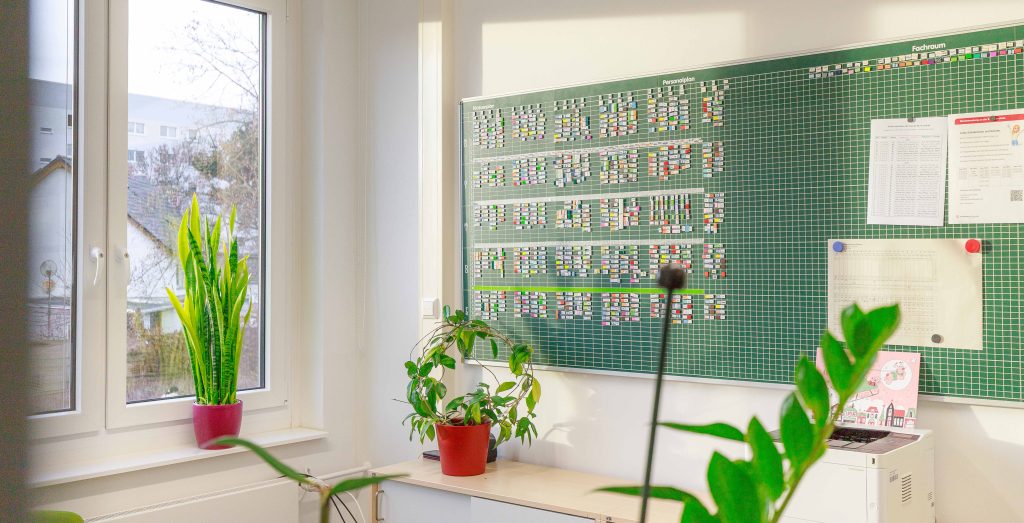 Ein sonnendurchflutetes Lehrerzimmer in Sachsen mit grünem Stundenplanbrett, mehreren Zimmerpflanzen, Unterlagen an der Wand und Fenstern am Rand.
