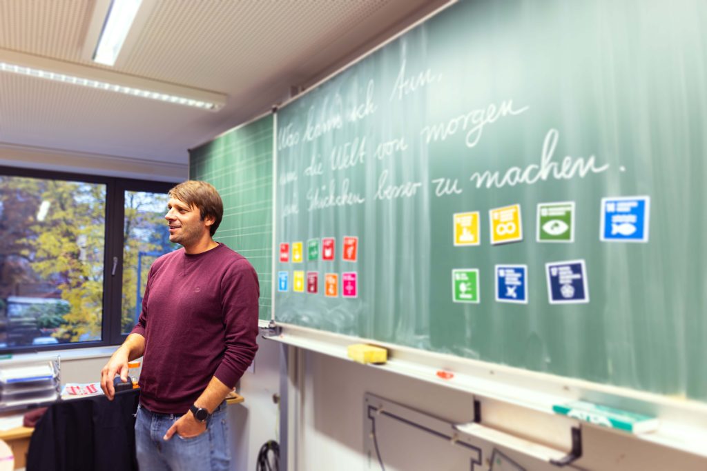 In einem Klassenraum steht eine Person mit bordeauxrotem Shirt vor einer grünen Tafel, auf der handschriftliche Notizen und farbige Symbole zu sehen sind.