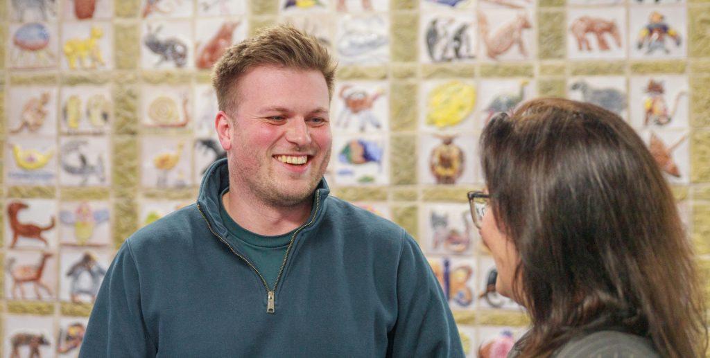 Es sind ein Mann links und eine Frau rechts vor einer Bildergalerie mit Zeichnungen von Tieren zu sehen. Der Mann schaut die Frau an, die Frau ist nur halb von hinten zu sehen,