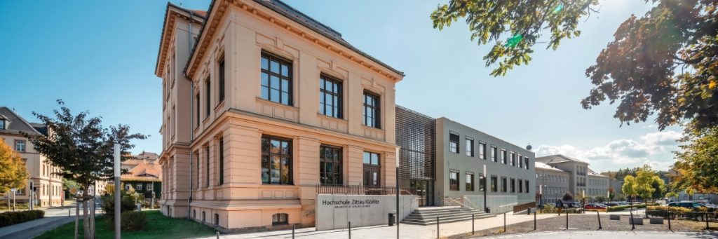 Stammhaus der Hochschule Zittau-Goerlitz mit peachfarbener Fassade, großen Fenstern und moderner Erweiterung am sonnigen Standort in Zittau.