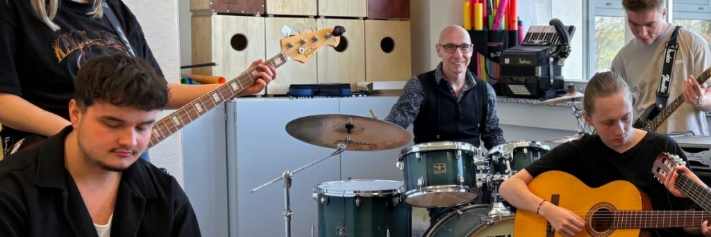 Michael Erblich lächelt im Proberaum vor blauem Schlagzeug, während Lernende mit Holzgitarre und Bassgitarre spielen und verschiedene Instrumente im Hintergrund stehen.