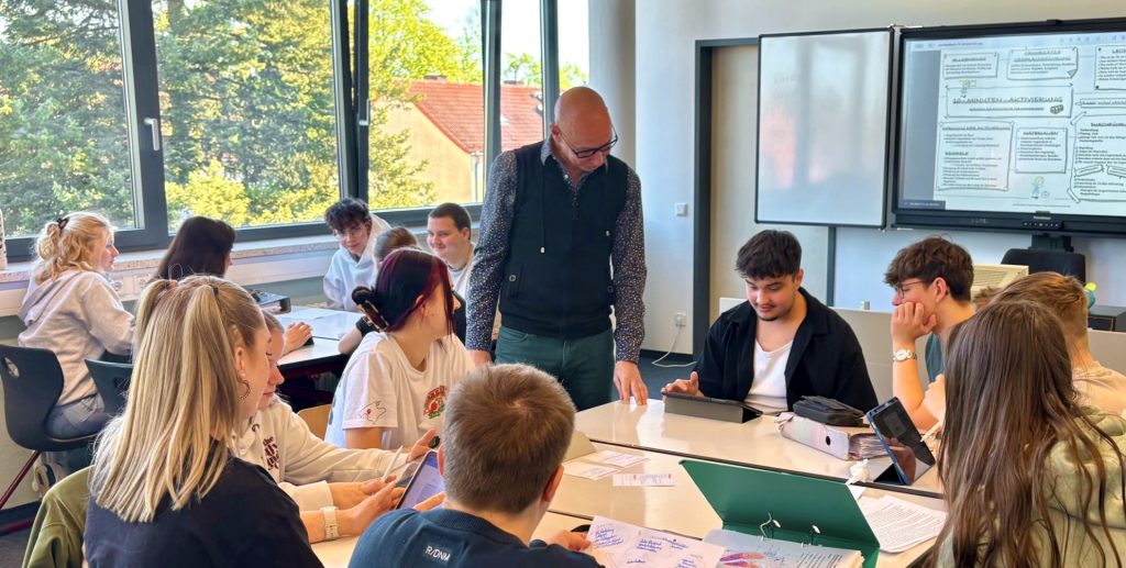 Ein Lehrer steht in einem Achtertisch in der Klasse und schaut einem Jungen auf das Tablet, auf dem die Schülerinnen und Schüler Aufgaben bearbeitet haben. Im Hintergrund ist ein Whitboard mit Tafelbild zu sehen, links ein weiterer Tisch mit Schülerinnen und Schülern, im Hintergrund links das Fenster mit Aussicht auf Bäume.