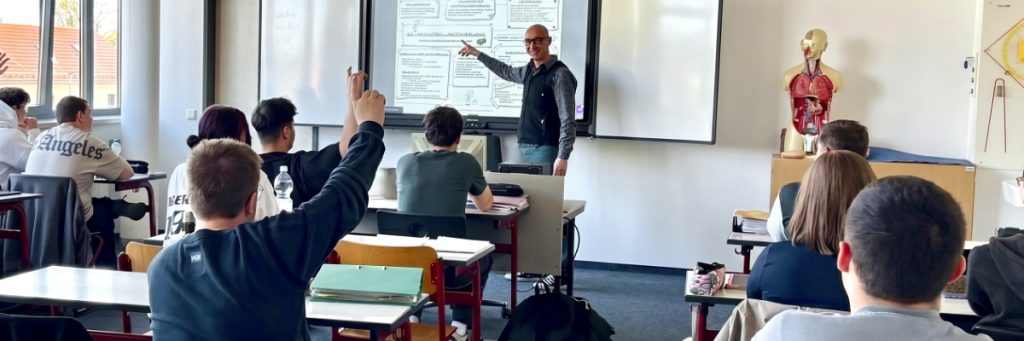 Michael Erblich unterrichtet im hellen Klassenzimmer vor Whiteboard und Display; Schüler hören zu, während Unterrichtspläne sichtbar sind.