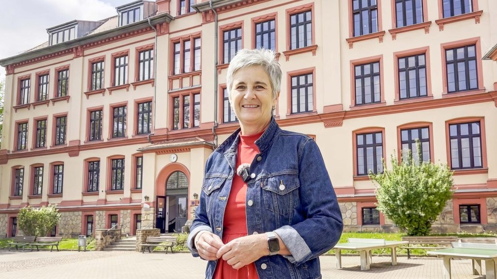 Eine lächelnde Frau mit Kurzhaarschnitt und Jeansjacke steht vor einem historischen Schulgebäude mit hohen Fenstern und rotem Putz.