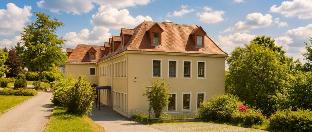 Das Berufsschulgebäude in Löbau steht inmitten grüner Anlagen mit gepflegten Wegen, beiger Fassade, rotem Dach und freundlicher Umgebung.