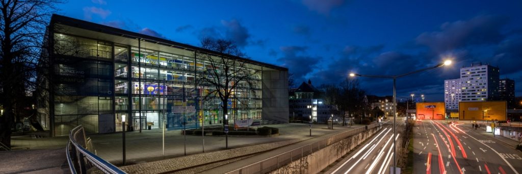 In der Nachtansicht ist links das erleuchtete moderne Gebäude des Hörsaalzentrums der TU Dresden mit seiner Glasfassade zu sehen. Rechts davon verläuft eine große Straße.