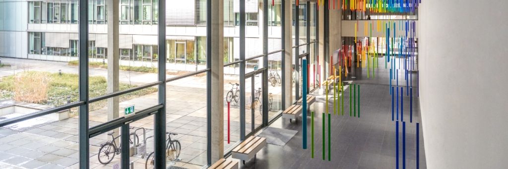 Es sind in einem Unigebäude der TU Dresden rechts in Regenbogenfarben herabhängende Stäbe als Kunst am Bau zu sehen. Links grenzt eine Glasfront das Gebäude zu einem Innenhof ab, in dem Fahrräder an Ständern geparkt sind.