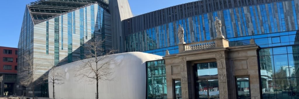 Links ist die blaue durchbrochene Glasfassade des Augusteums an der Uni Leipzig zu sehen, mittig ein moderner Betonvorbau mit abgerundeten Kanten, daneben ein historisches Portal vor der Glasfront, auf dem zwei Figuren-Skulpturen stehen.