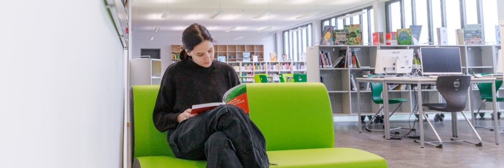 Eine Schülerin sitzt in einer Schulbibliothek auf einer Bank und liest ein Buch.