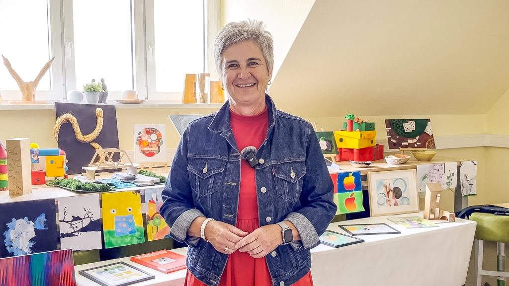 Eine lächelnde Frau im roten Kleid und Jeansjacke steht vor einer Ausstellung farbenfroher Schülerarbeiten.