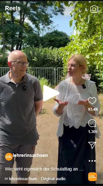 Ein älterer Mann mit Brille und eine blonde Frau sprechen auf einem sonnigen Weg vor einem Zaun unter grünen Bäumen.