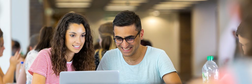 Zwei Studierende arbeiten lächelnd gemeinsam an einem Laptop in einer belebten Hochschulumgebung am Campus Augustusplatz der Universität Leipzig.