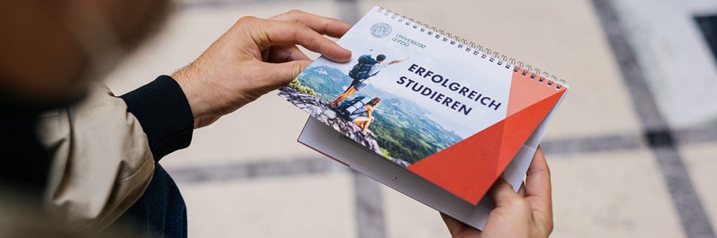 Ein Mensch hält einen Studienratgeber mit dem Titel „Erfolgreich studieren“ der Universität Leipzig in den Händen. Auf dem Foto auf der Brsochüre sind zwei junge Menschen vor einem Landschaftspanorama zu sehen.