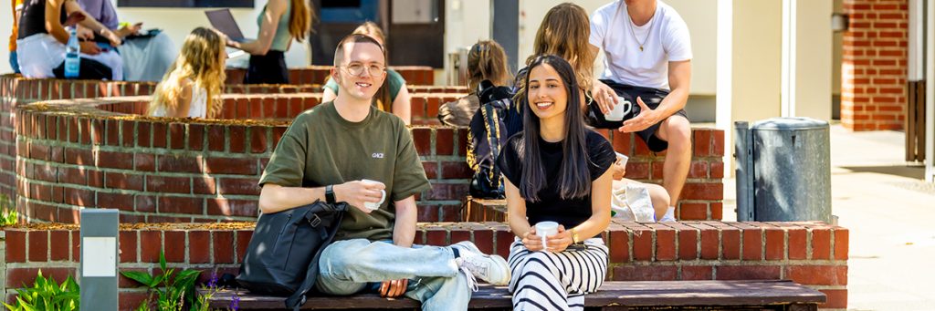 Zwei Studentinnen sitzen lachend auf einer Backsteinmauer vor einem belebten Campus mit weiteren jungen Menschen.