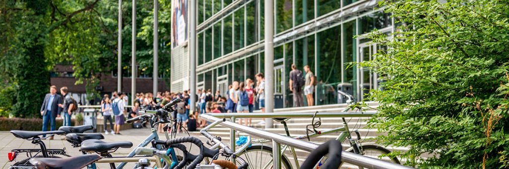 Vor einem Hochschulgebäude der TU Dresden mit großer Glasfront stehen Fahrräder, während Menschen am Eingang und auf den Wegen unterwegs sind.