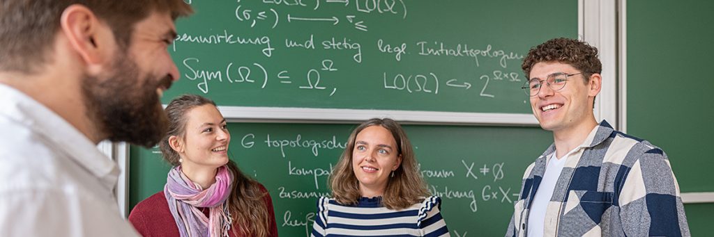 Vier Studierende sind vor einer grünen Kreidetafel mit mathematischen Zeichen und Notizen im Austausch miteinander