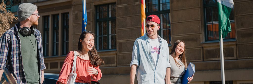 Vier Studierende mit Studienunterlagen unter den Armen sind vor einem Gebäude der Hochschule Zittau/Görlitz in Bewegung